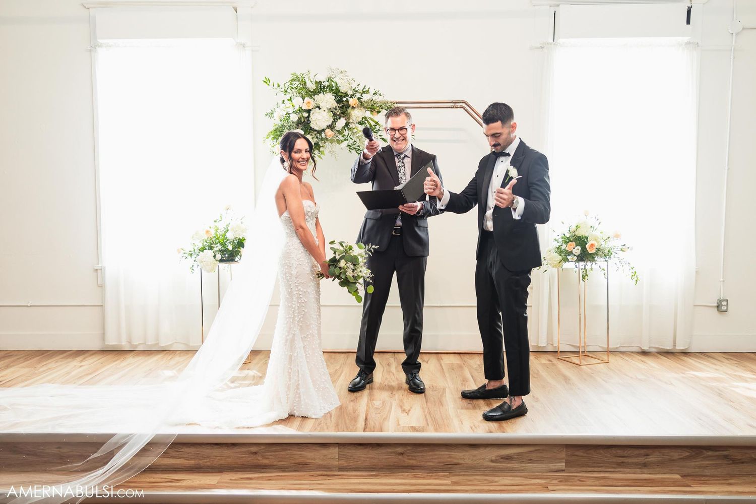 Wedding ceremony in bright indoor venue with white backdrop and floral arrangements.