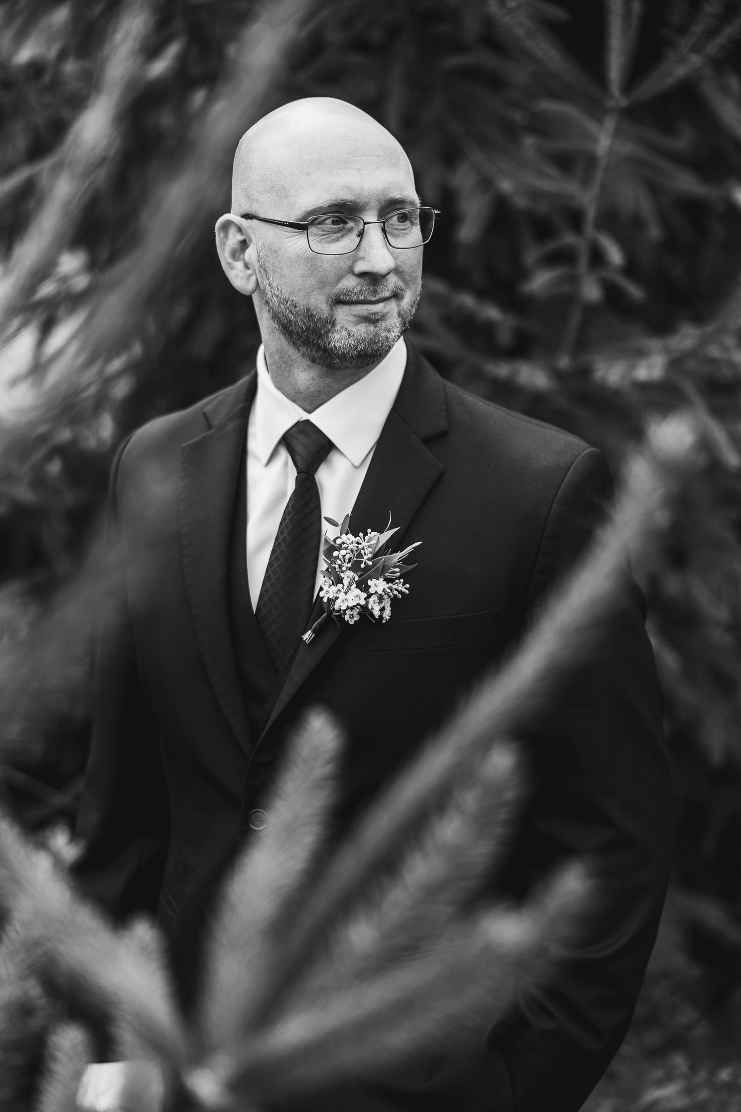 Close-up portrait of a groom in black suit and glasses smiling during an outdoor wedding ceremony.