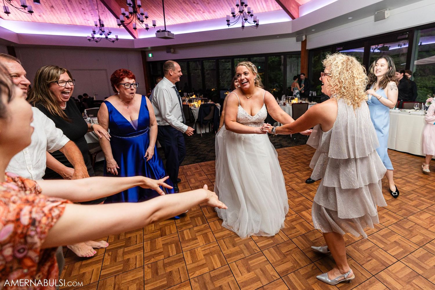 Wedding guests dancing in a circle on a parquet floor during a lively reception celebration.