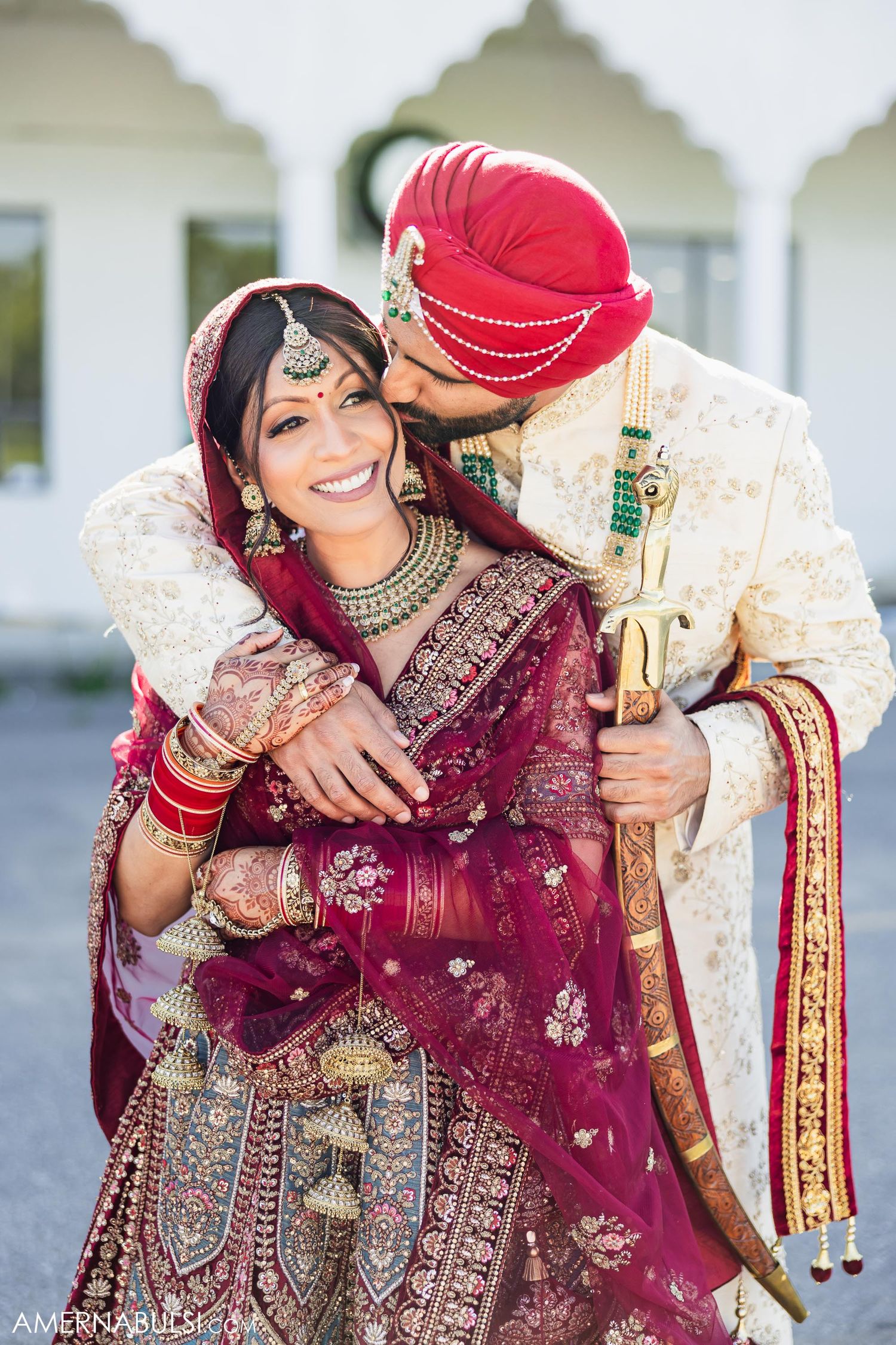 Traditional Indian wedding couple embracing in ornate attire against white architectural backdrop.