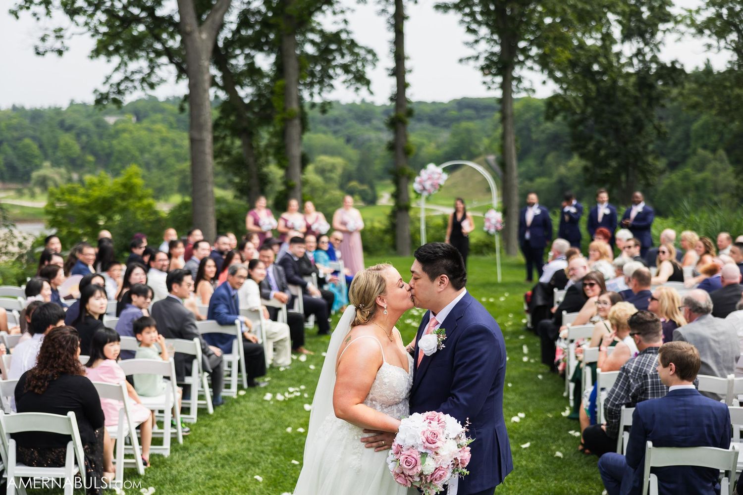 Outdoor wedding ceremony with couple kissing under floral arch surrounded by seated guests.