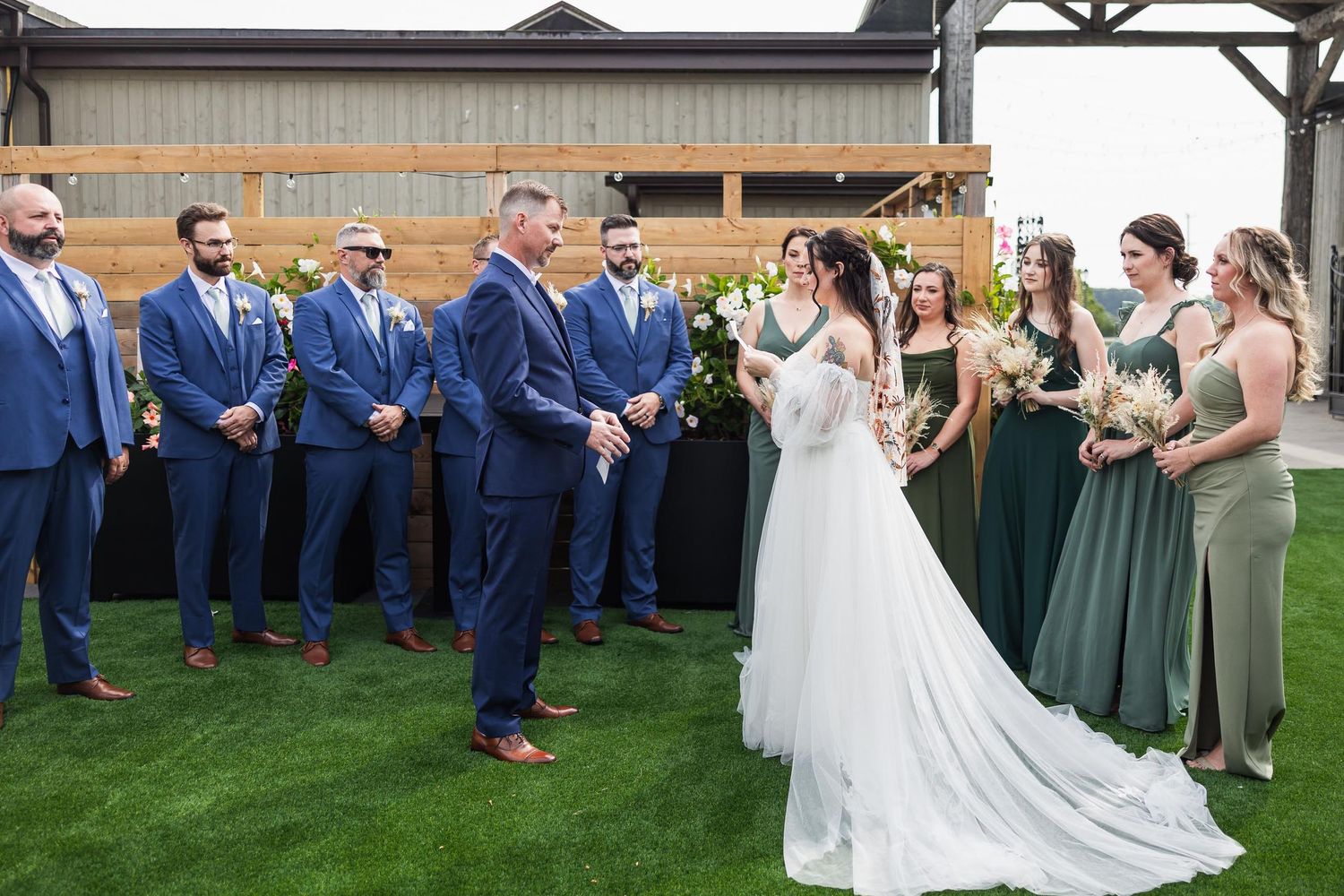 Wedding ceremony with attendants in navy suits and green dresses on outdoor lawn.