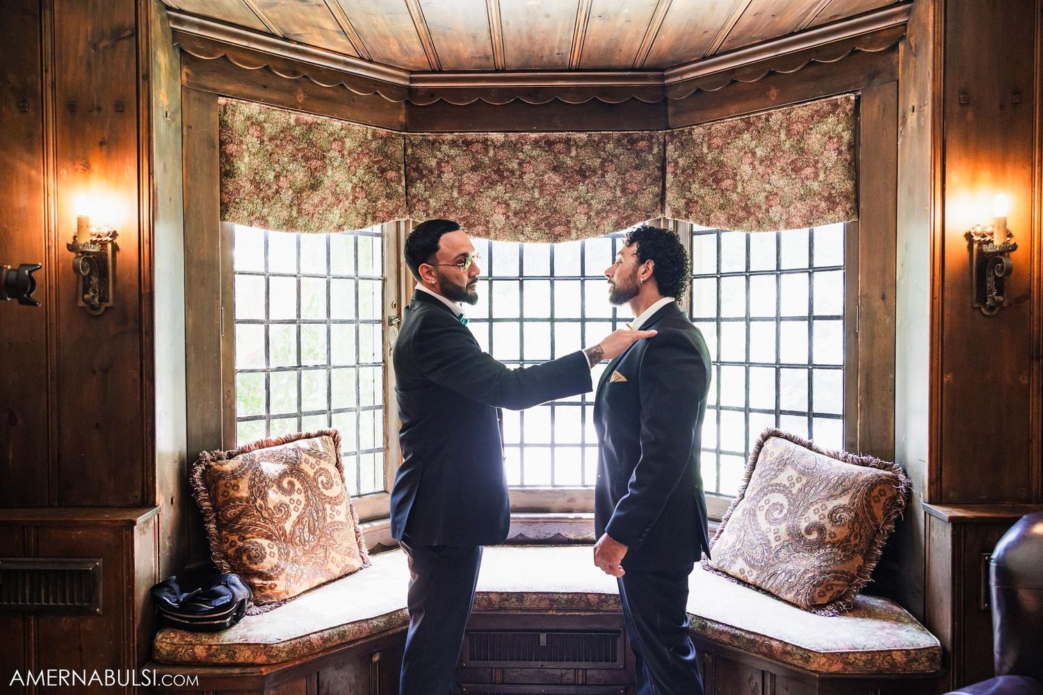 Two people in formal suits adjusting each other's ties by a window in an elegant room.