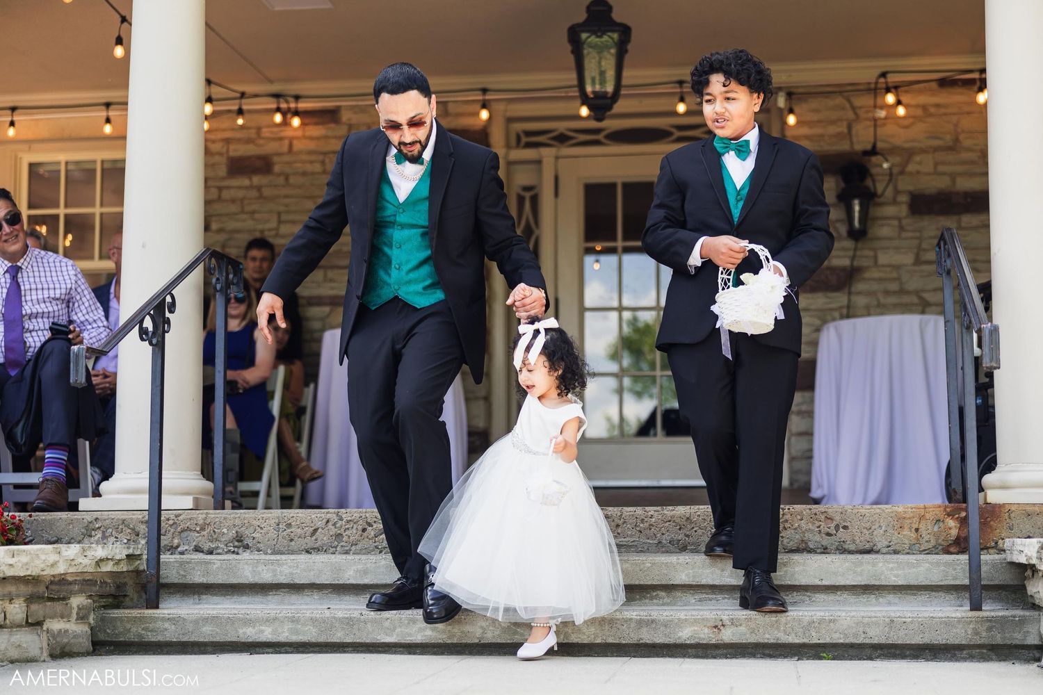 A flower girl being escorted down stone steps by groomsmen in teal and black suits.
