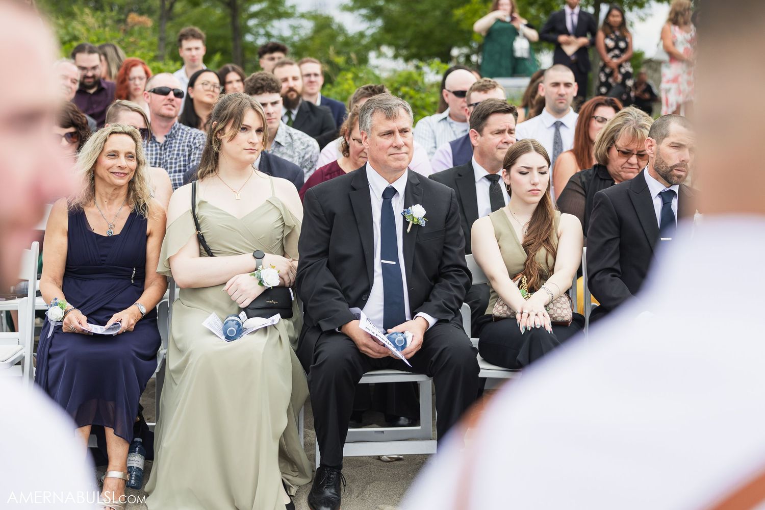 Wedding guests seated in white chairs watch an outdoor ceremony by the water.