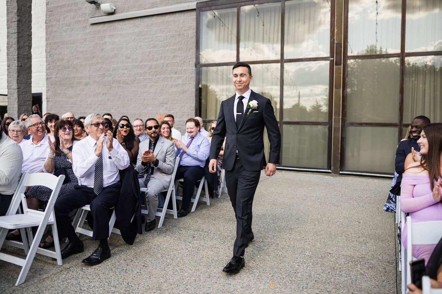 Groom walks down the aisle in a dark suit with boutonniere as wedding guests applaud at an outdoor ceremony.