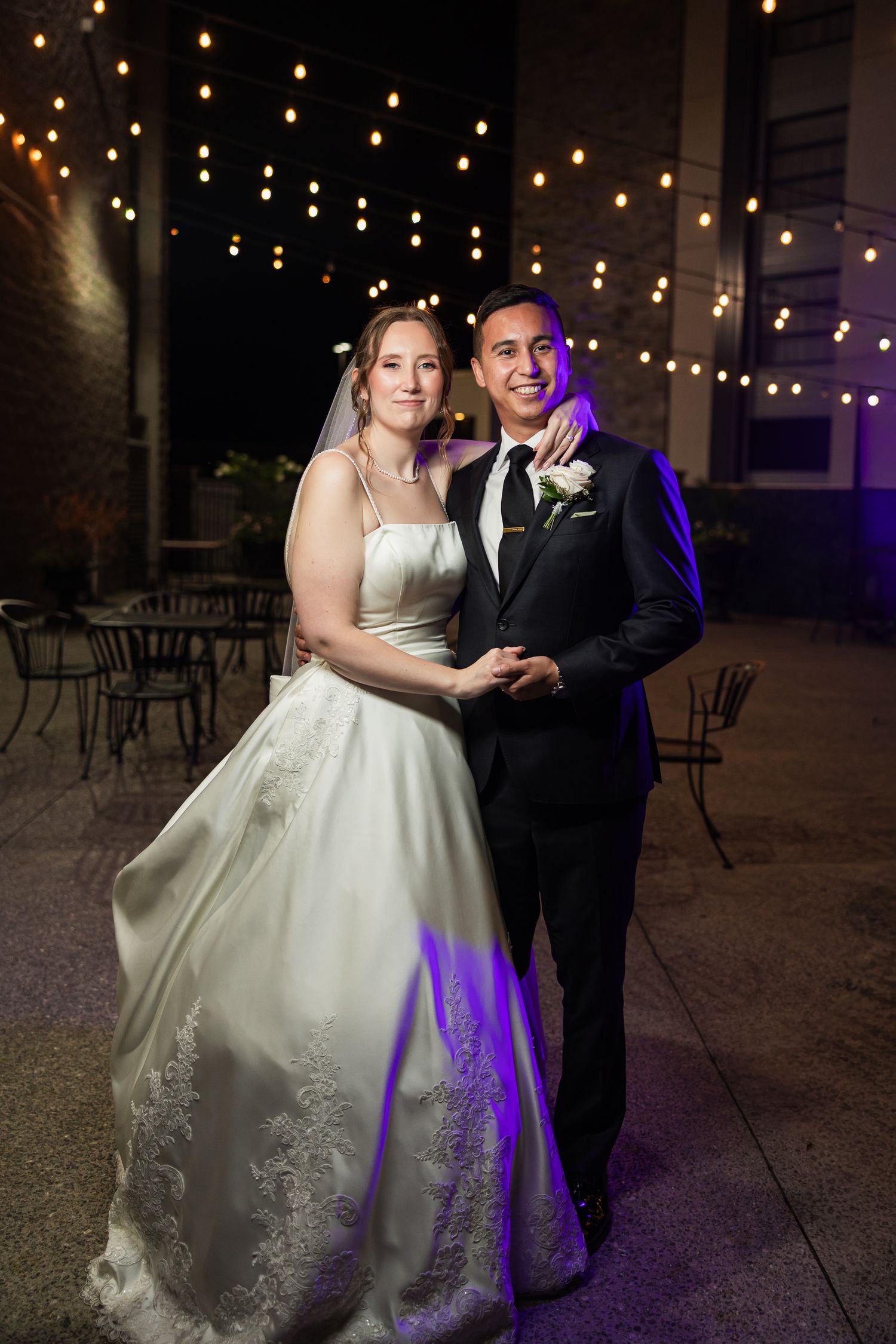 Bride in white lace gown and groom in black tuxedo pose outdoors at night under string lights.