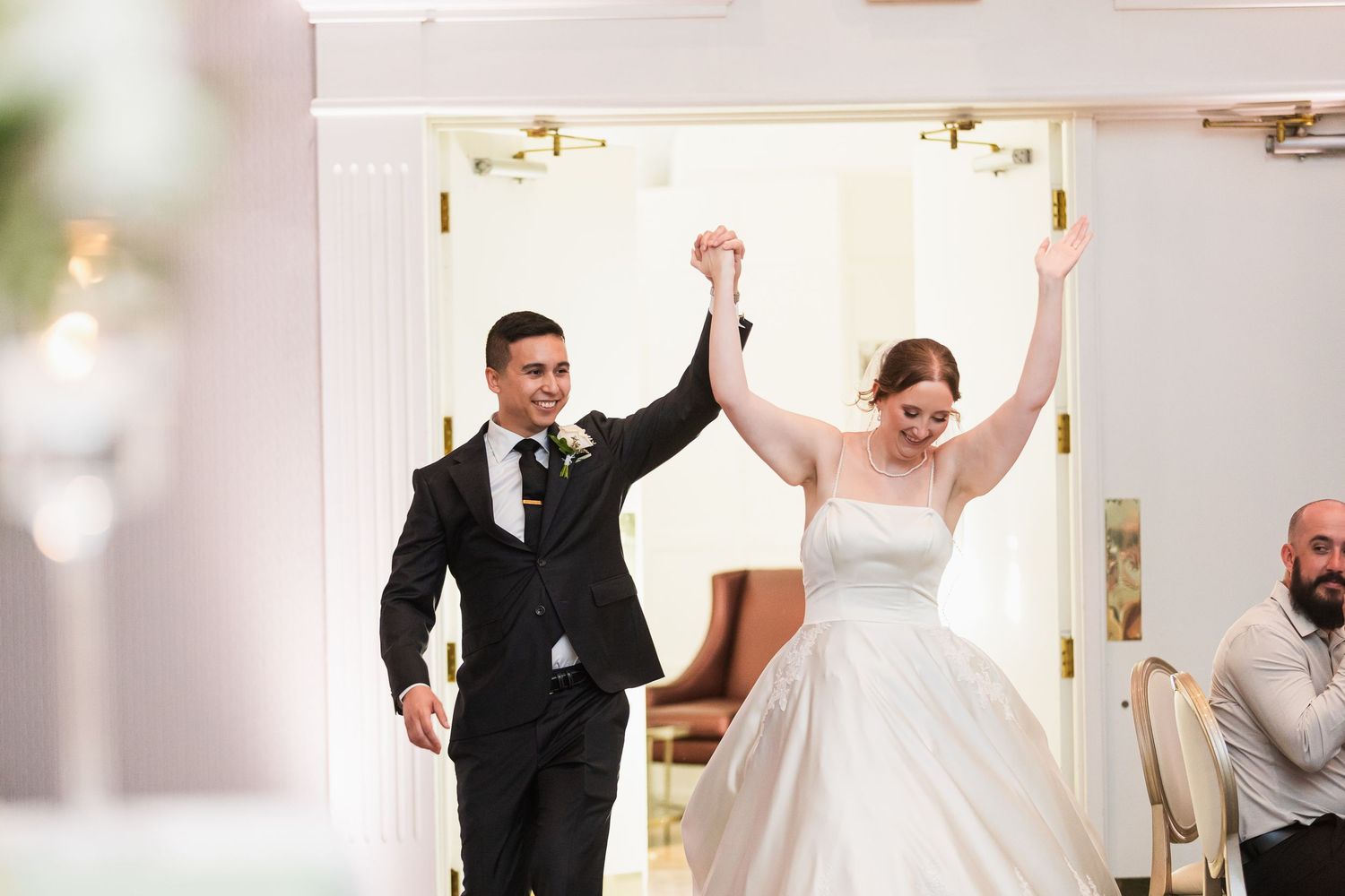 Bride and groom make grand entrance at wedding reception, raising hands in celebration in elegant white venue.