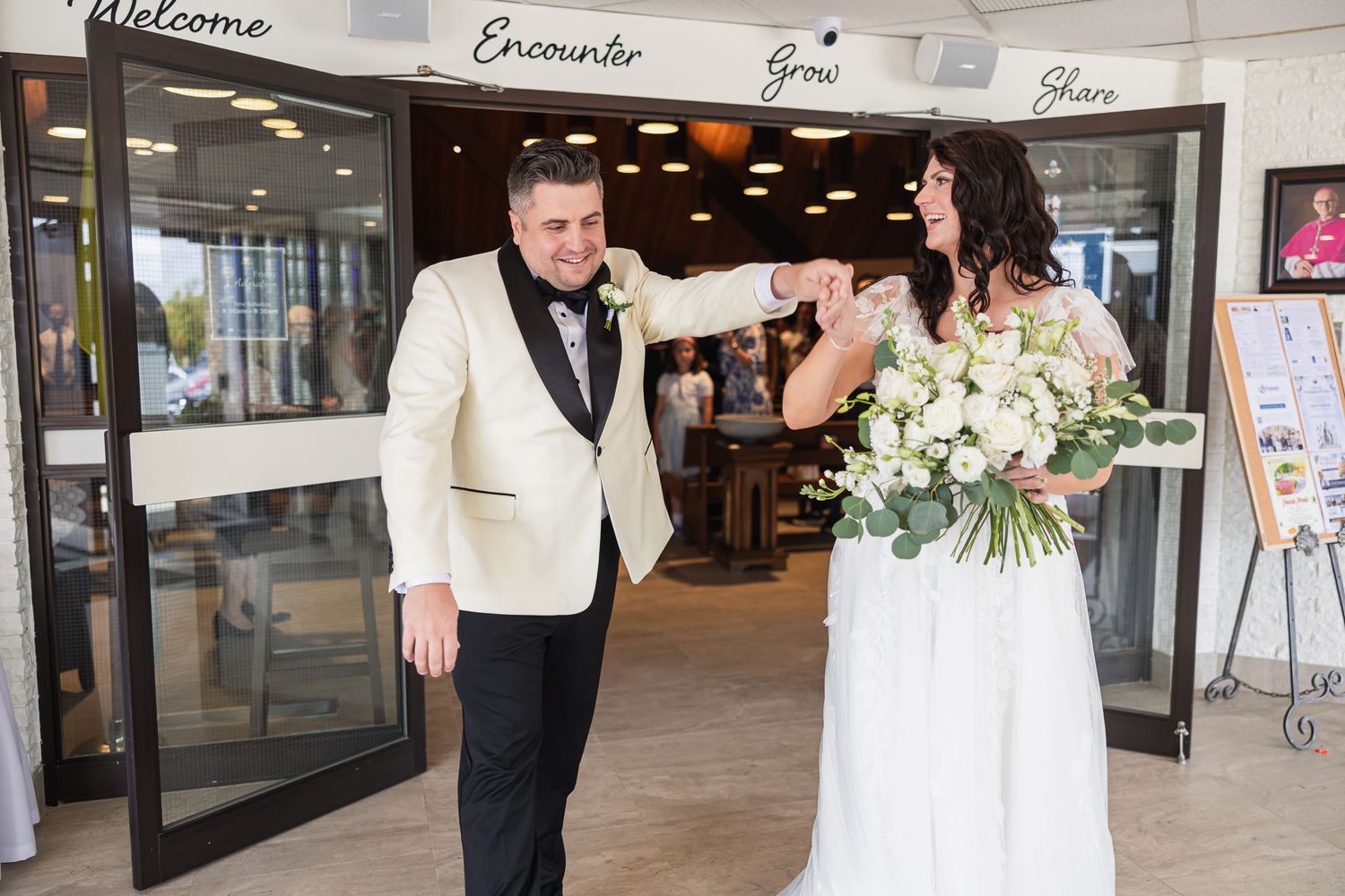 Bride and groom share a joyful fist bump outside wedding venue entrance, bride holding white floral bouquet.