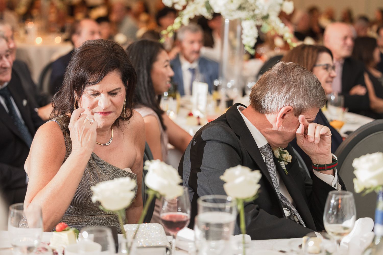 Emotional wedding guests wiping tears at a reception dinner table adorned with white roses and candles.