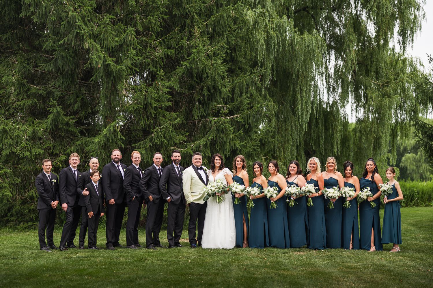 Wedding party with groomsmen in black suits and bridesmaids in teal dresses posing outdoors.