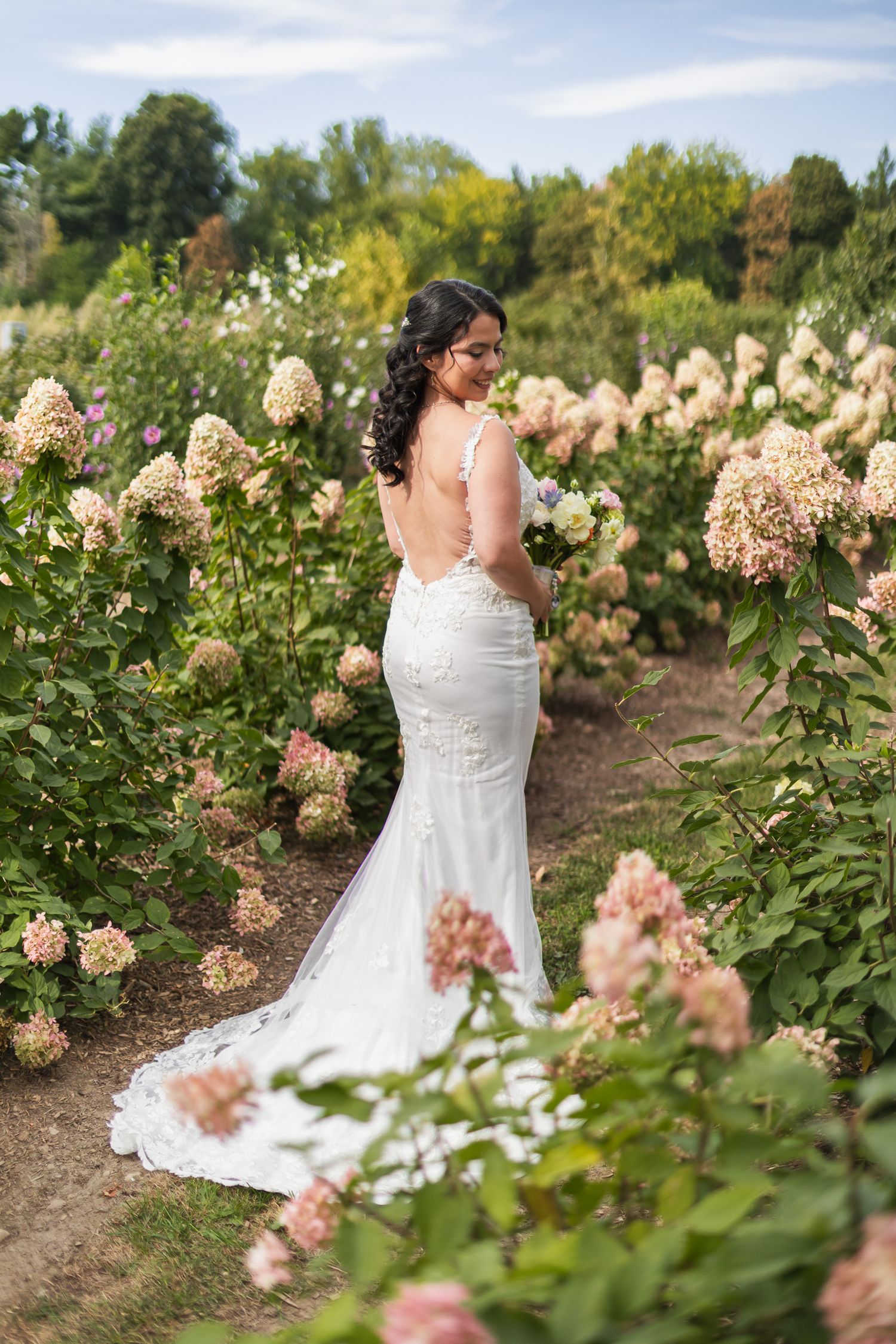 Bride in backless lace mermaid gown holding bouquet among blooming hydrangeas in garden.