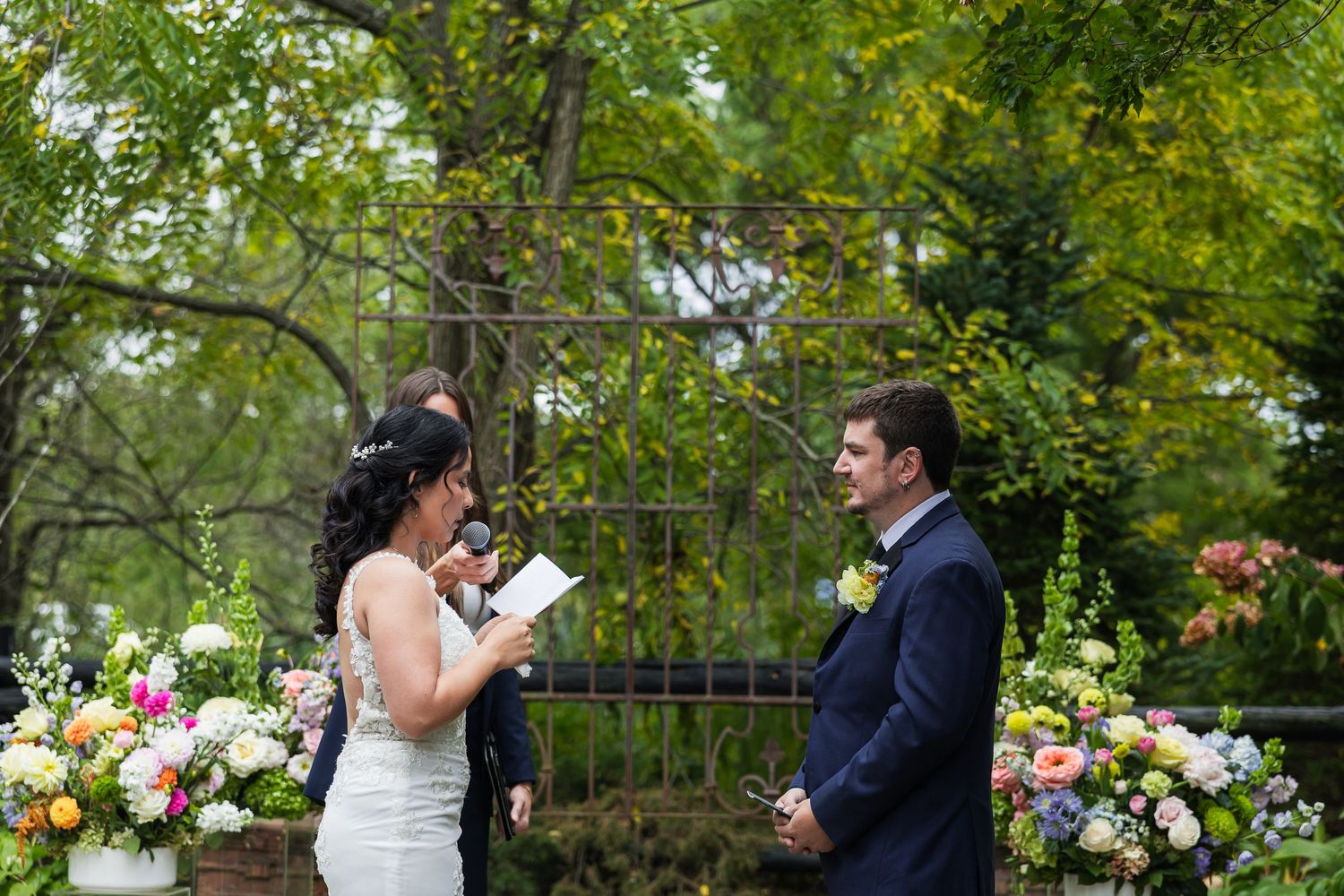Bride reads vows to groom at outdoor wedding ceremony surrounded by colorful floral arrangements and lush greenery.