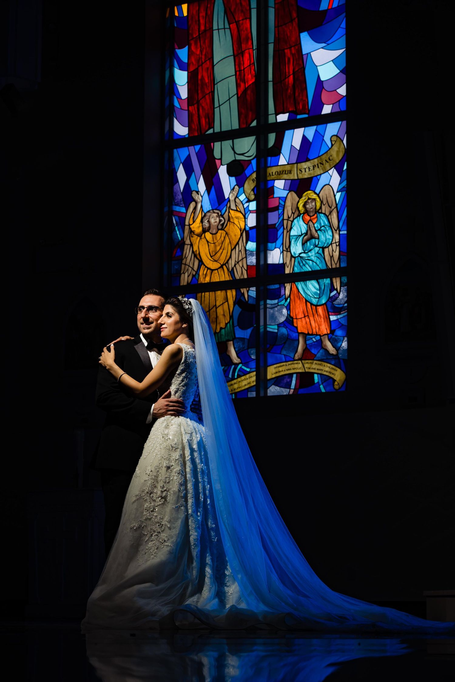 Elegant couple poses in front of dramatic stained glass windows in a dark church interior.