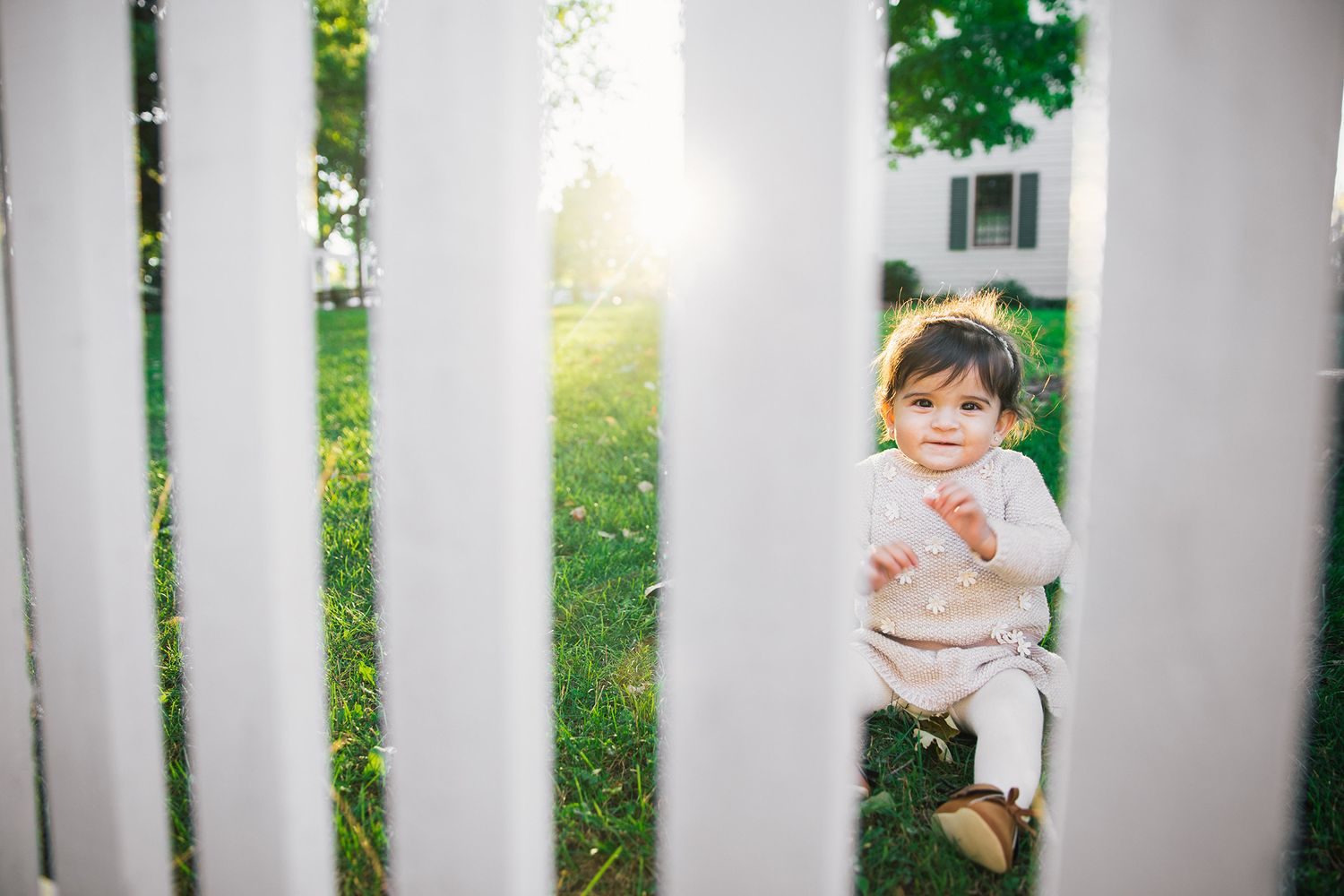 Small child peeks through white fence slats in garden with sunlight filtering through trees in background.