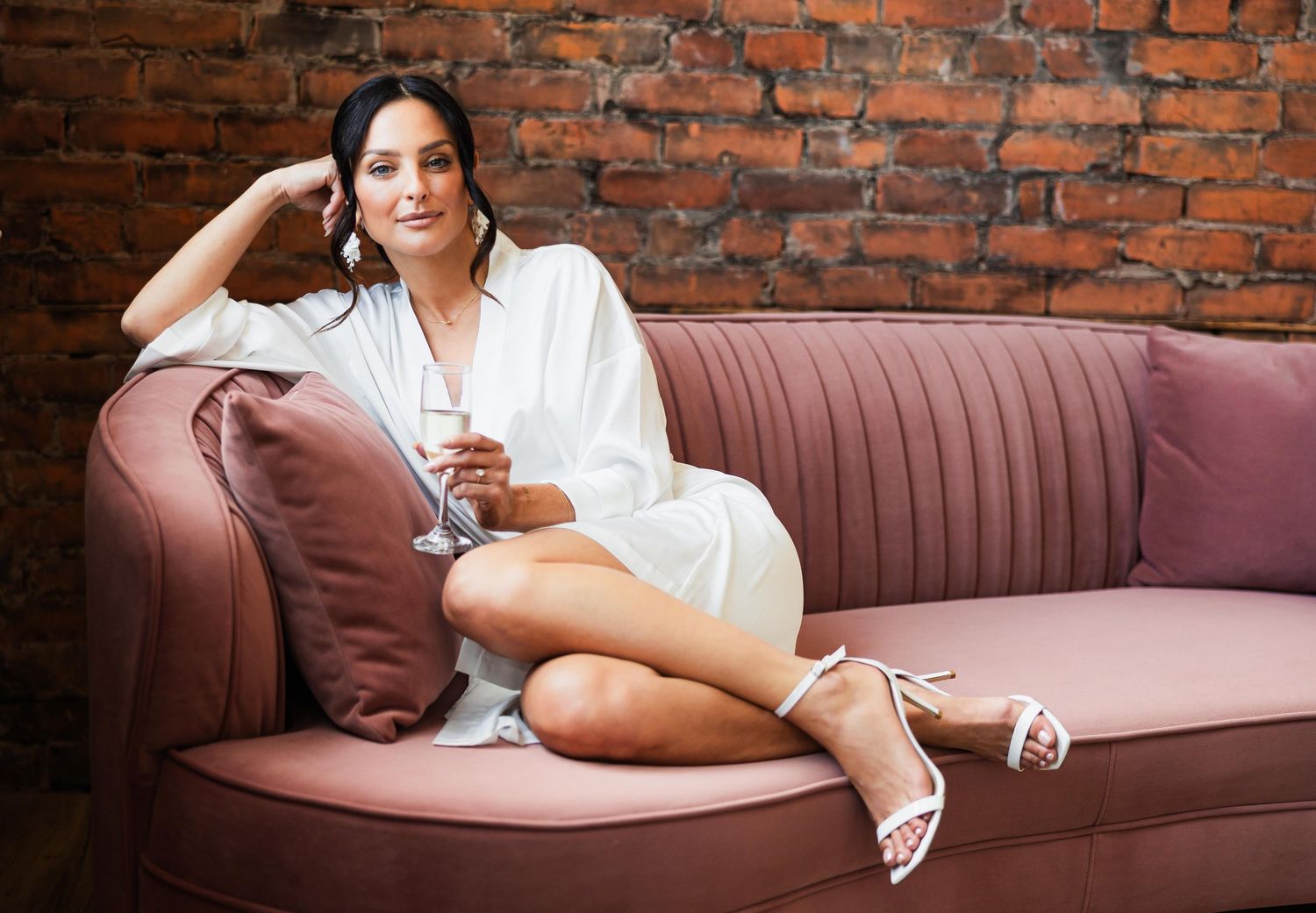 Bride relaxes on pink vintage couch against exposed brick wall.