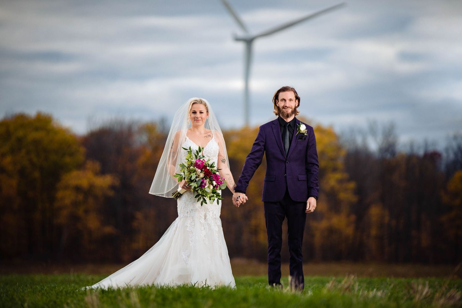 A couple poses in formal wedding attire with a wind turbine visible in the background.