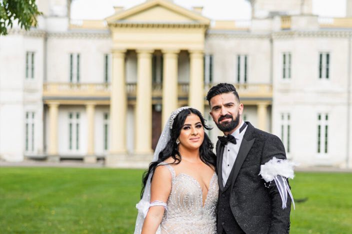 Elegant couple poses in front of stately mansion with classical columns and architecture.