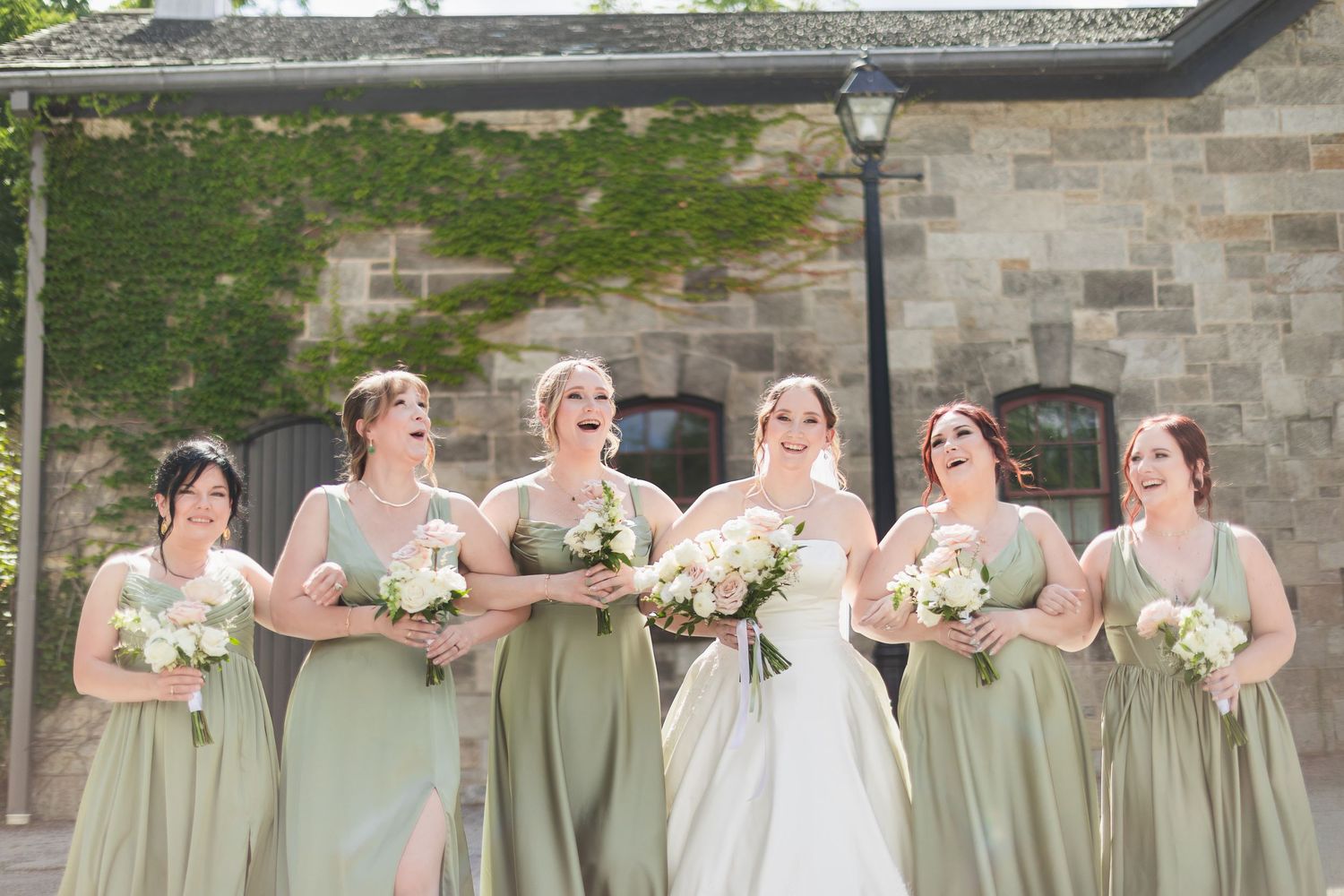 Bridesmaids in sage green gowns laugh with bride in white outside stone building.