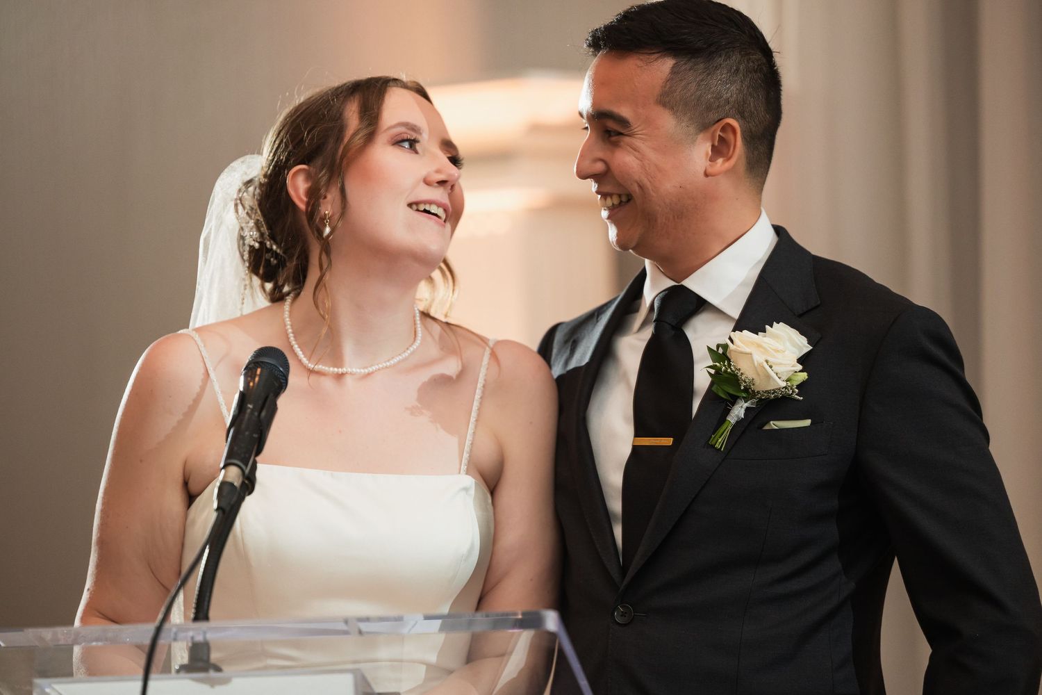 Bride and groom smiling at each other at wedding altar, bride in white dress with pearl necklace, groom in black suit.
