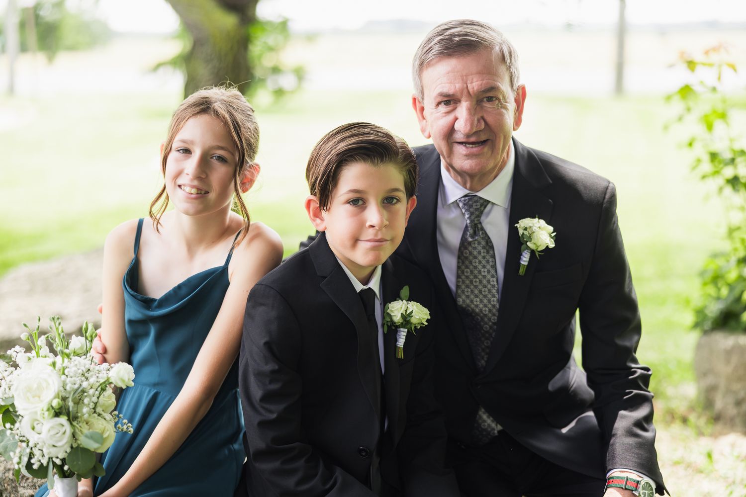 Older man in suit with boutonniere posing with two young children in formal attire at a wedding.