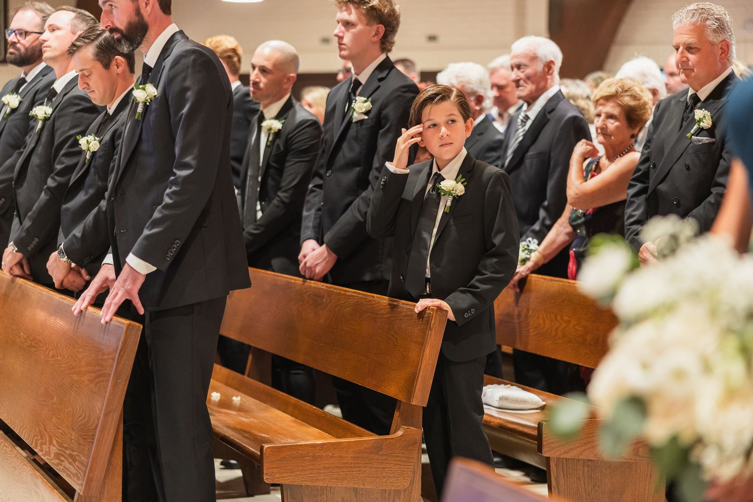 Groomsmen in black suits with white boutonnieres stand in church pews during a formal wedding ceremony.