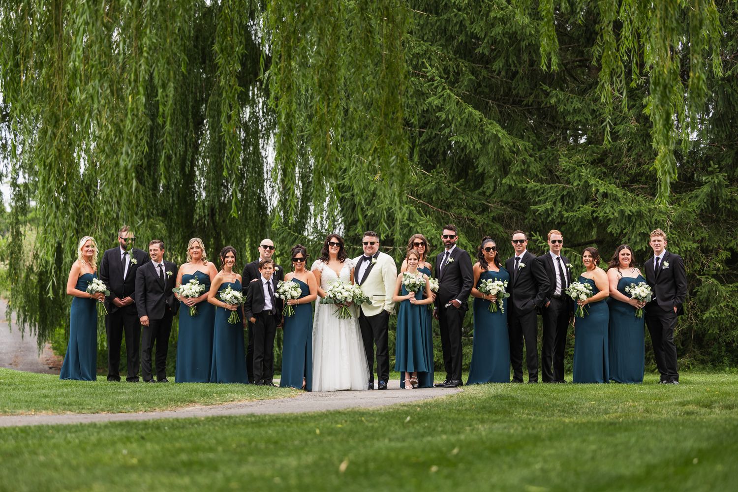Large wedding party poses together under weeping willow trees, bridesmaids in teal gowns, groomsmen in black suits.