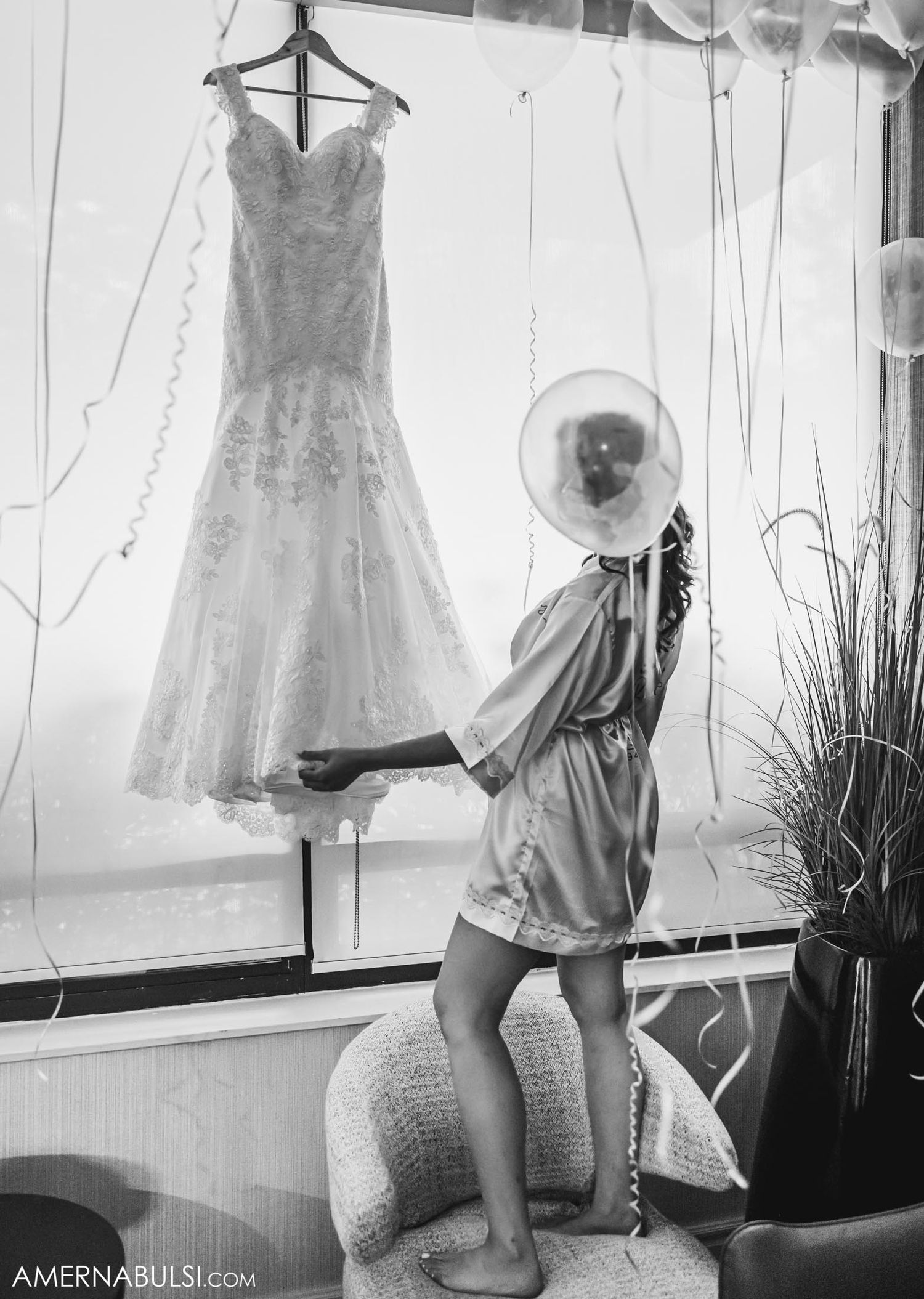 Artistic black and white photo of a wedding dress hanging as someone poses with flash photography equipment.