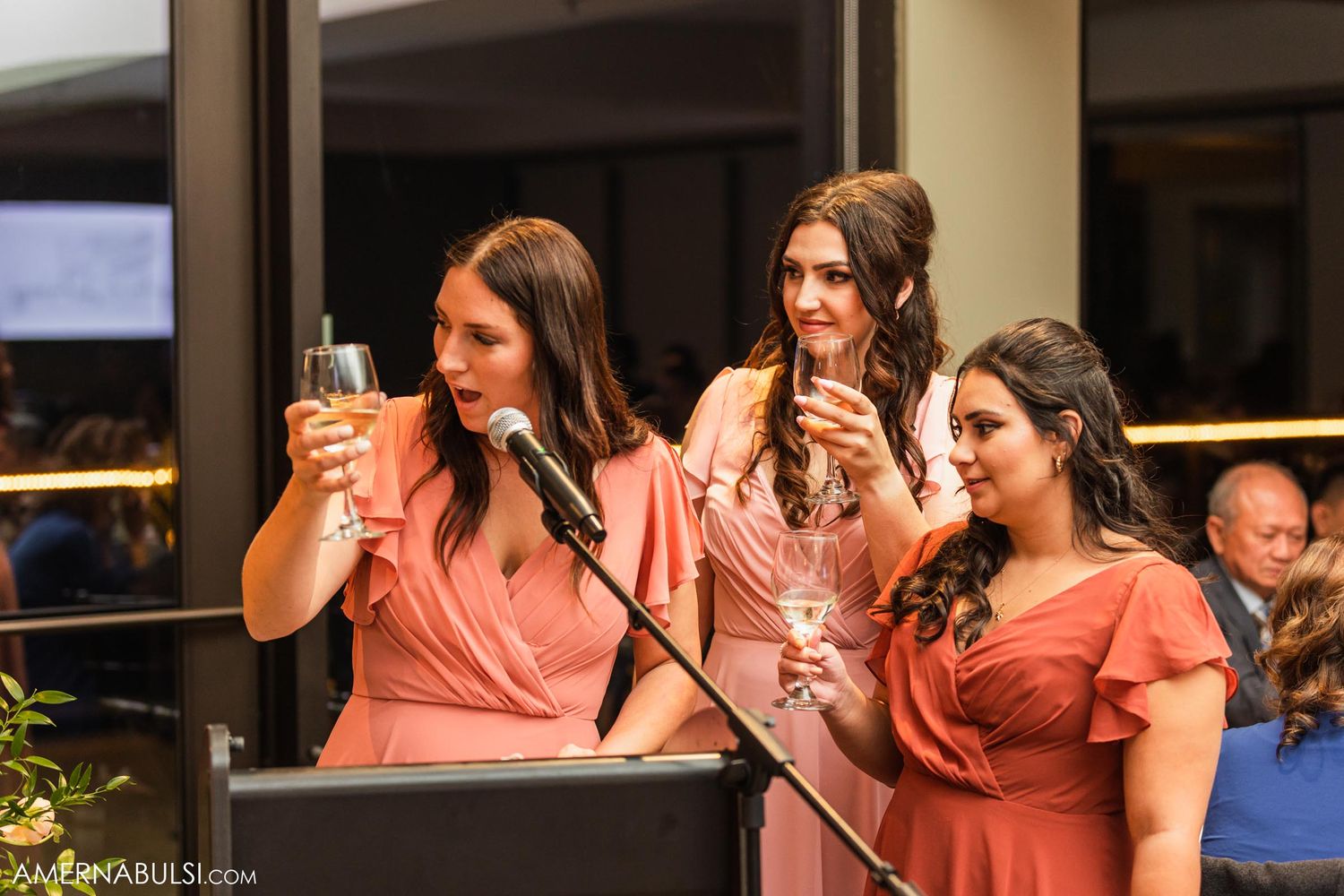 Bridesmaids in coral dresses giving toasts during wedding reception speeches.