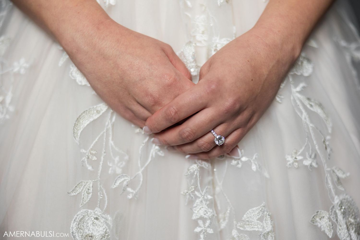 Close-up of hands resting on white lace wedding dress fabric with engagement ring.