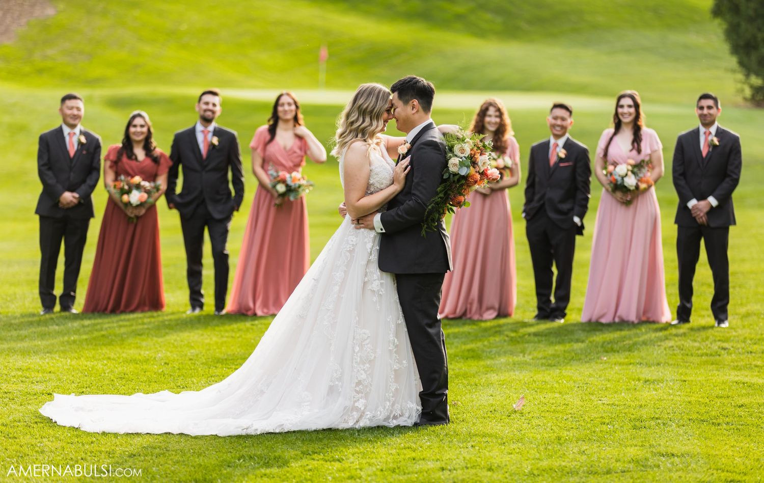 Wedding party lined up on green hillside while newlyweds share romantic moment.
