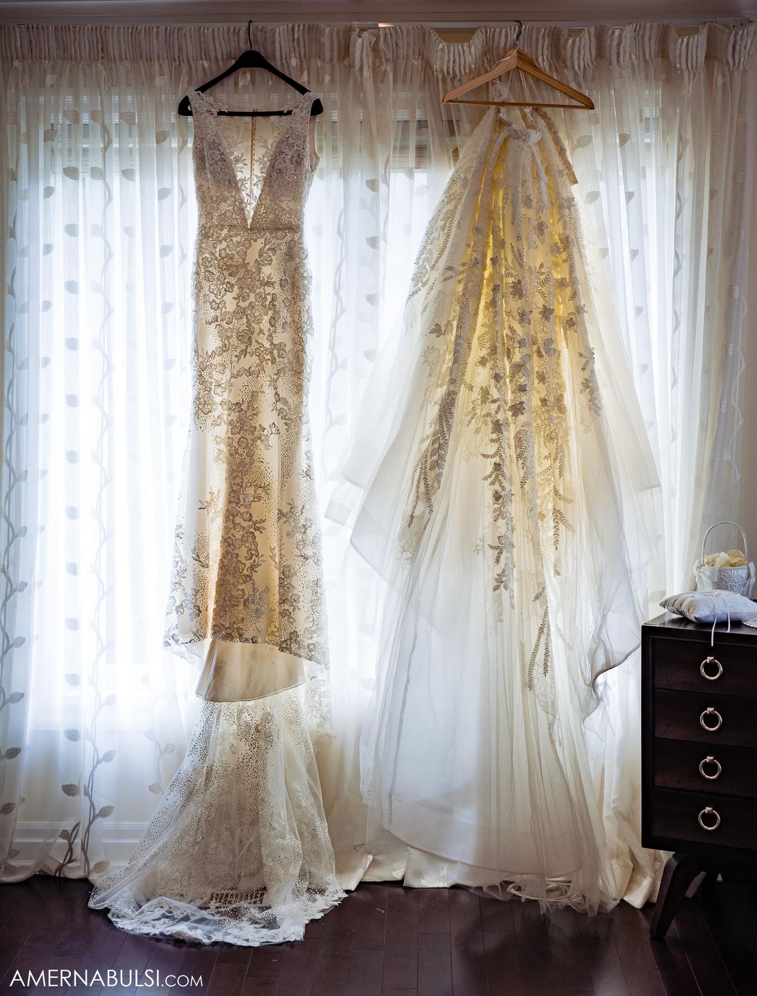 Two ornate wedding dresses hanging side by side against sheer white curtains in natural light.