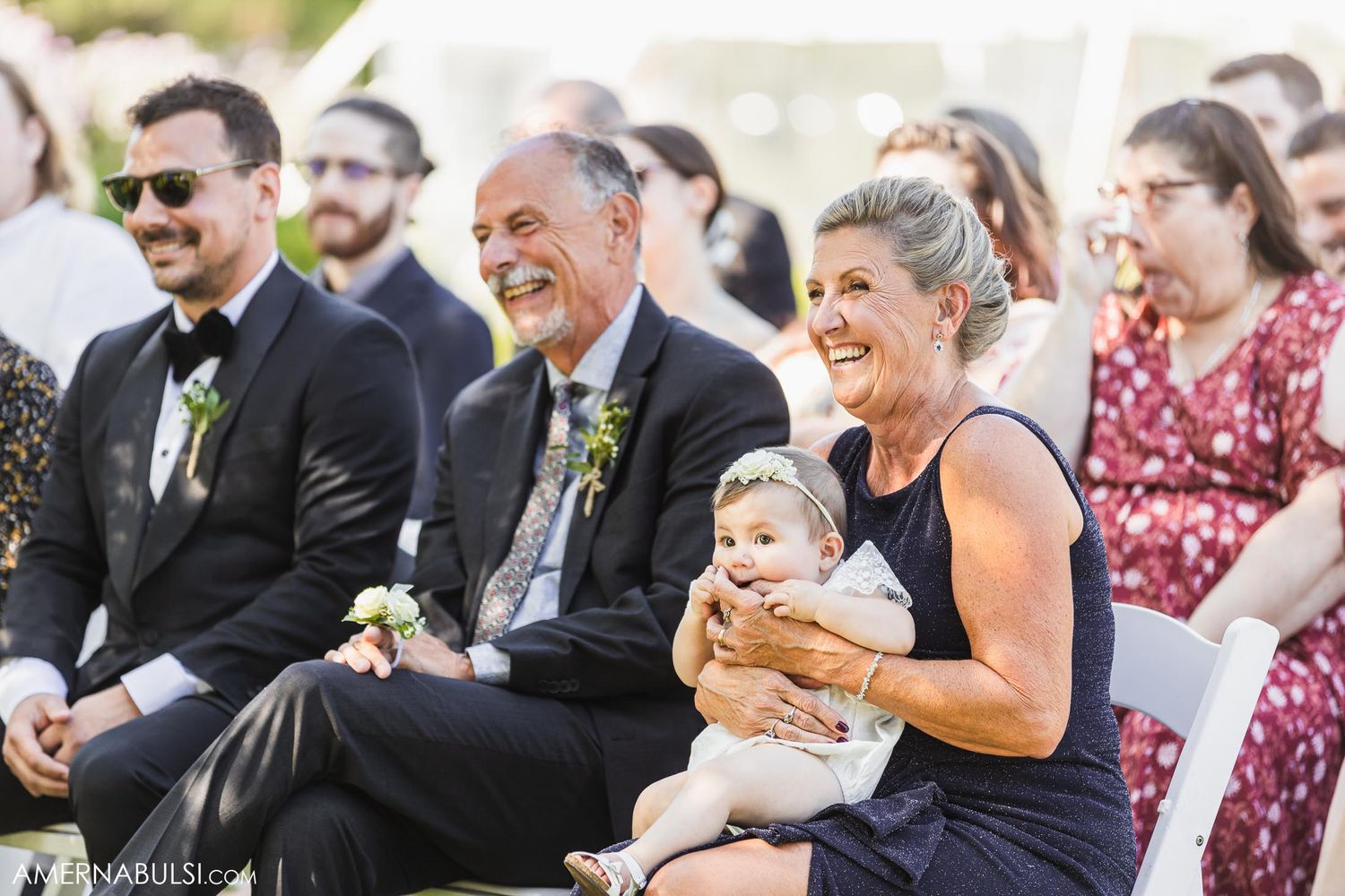 Wedding guests smile and laugh while watching the ceremony from their seats on a bright afternoon.