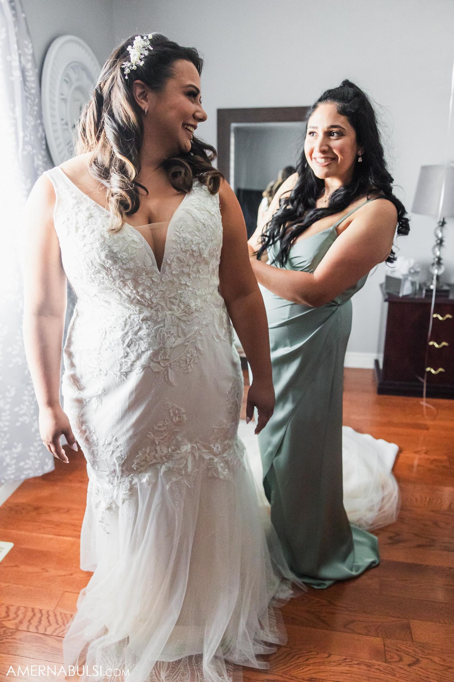 Bride in lace wedding gown stands with bridesmaid in sage green dress during fitting.