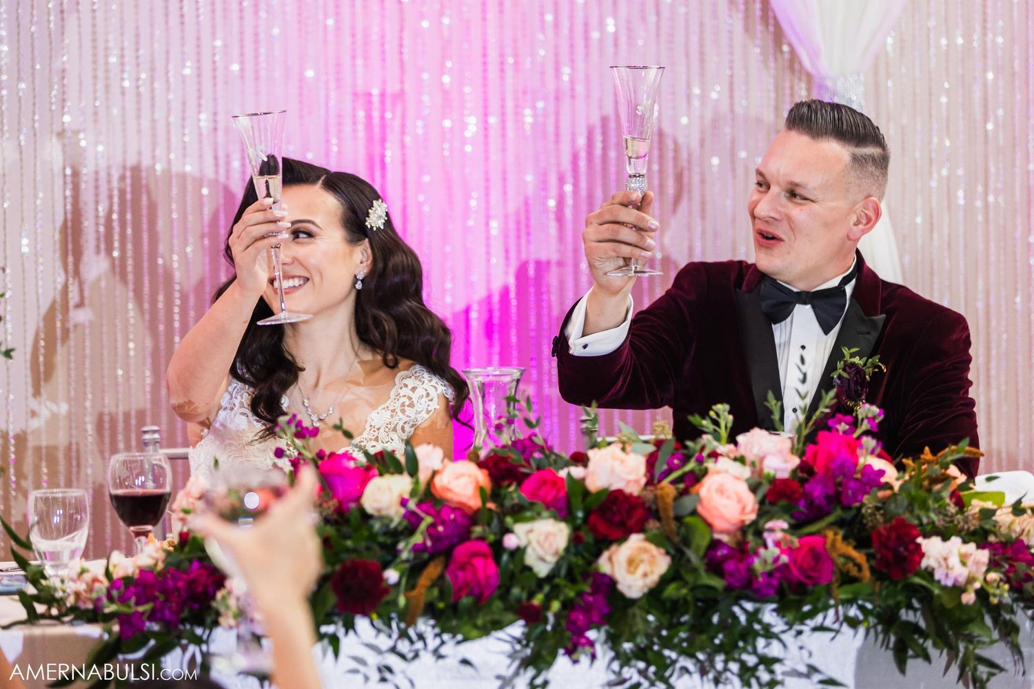 Wedding couple shares a champagne toast at their reception with pink decor and floral centerpieces.