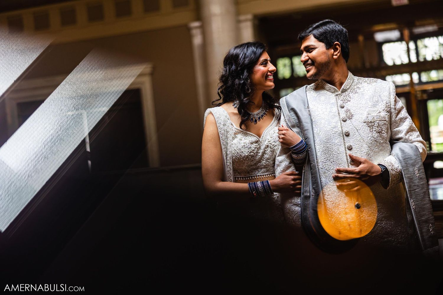 Couple in white and silver Indian wedding attire share a moment in atmospheric lighting.