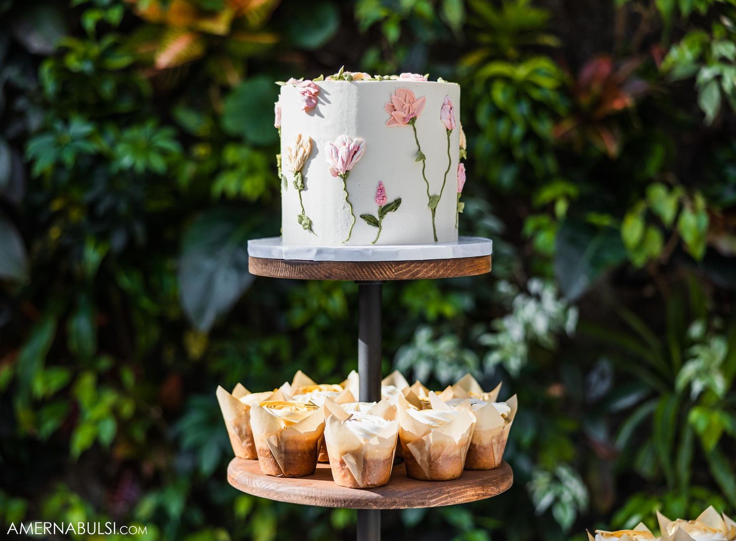 Two-tiered white cake with delicate pink floral design displayed with cupcakes on rustic wood stand.