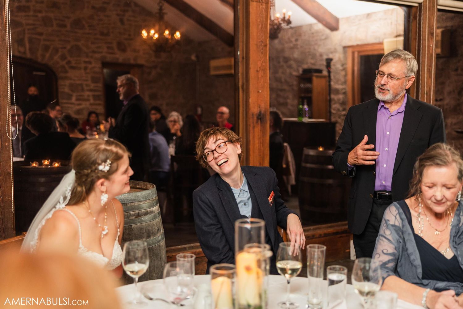 Wedding reception guests enjoying conversation and drinks at a table in a rustic venue.