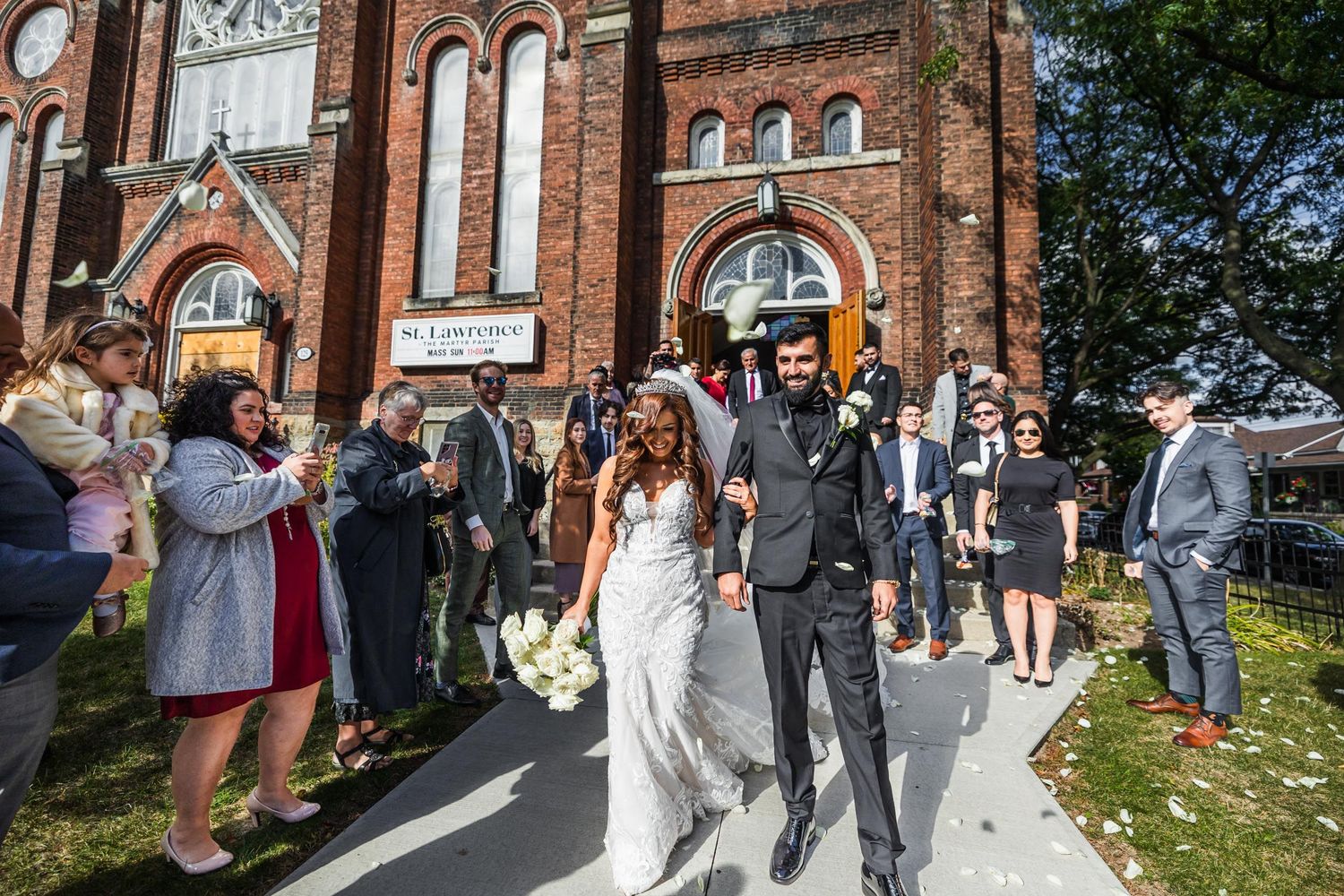 Newlyweds exit historic brick church while guests celebrate with flower petals on sunny day.