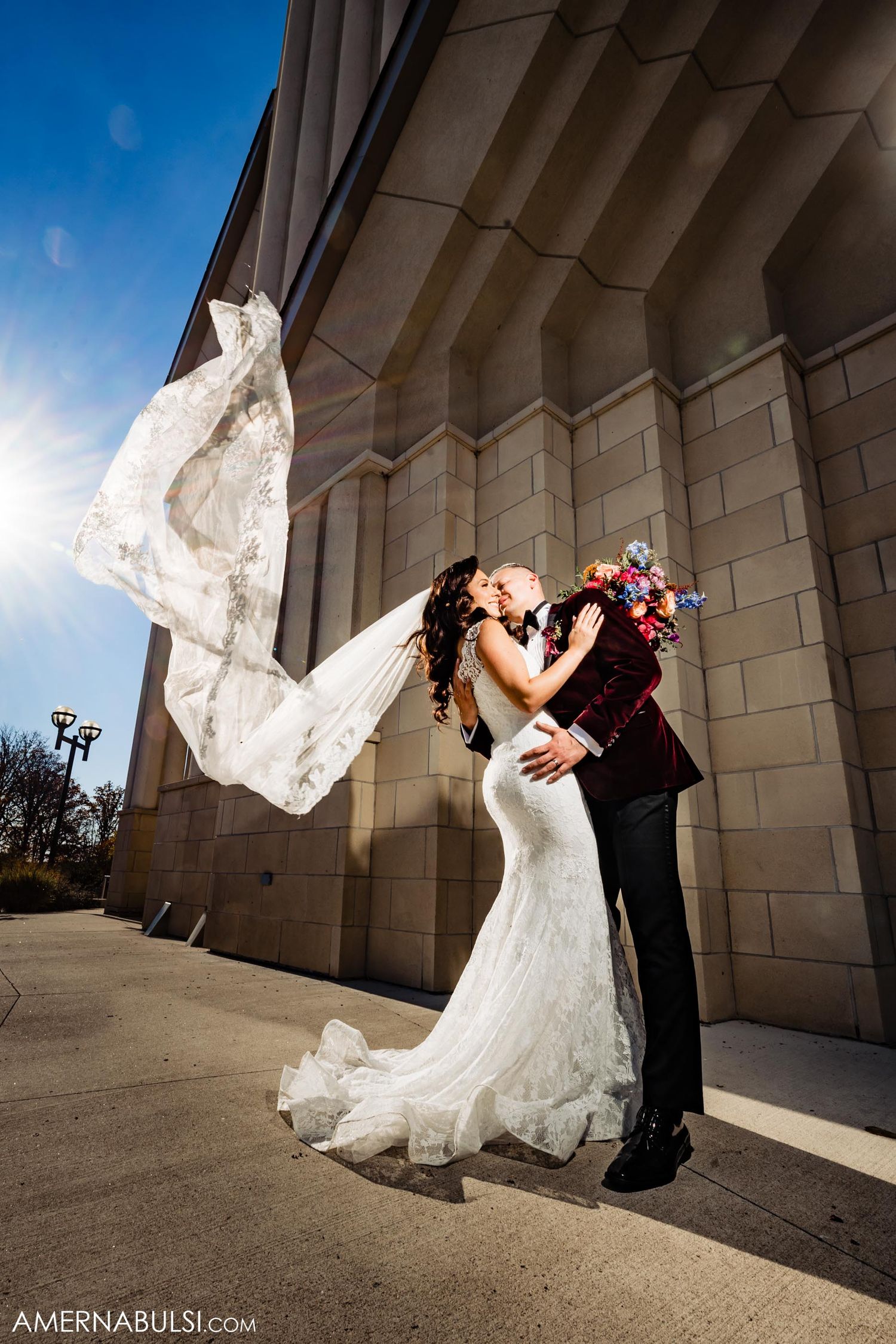 Dramatic wedding photo against modern building with flowing veil caught in wind.