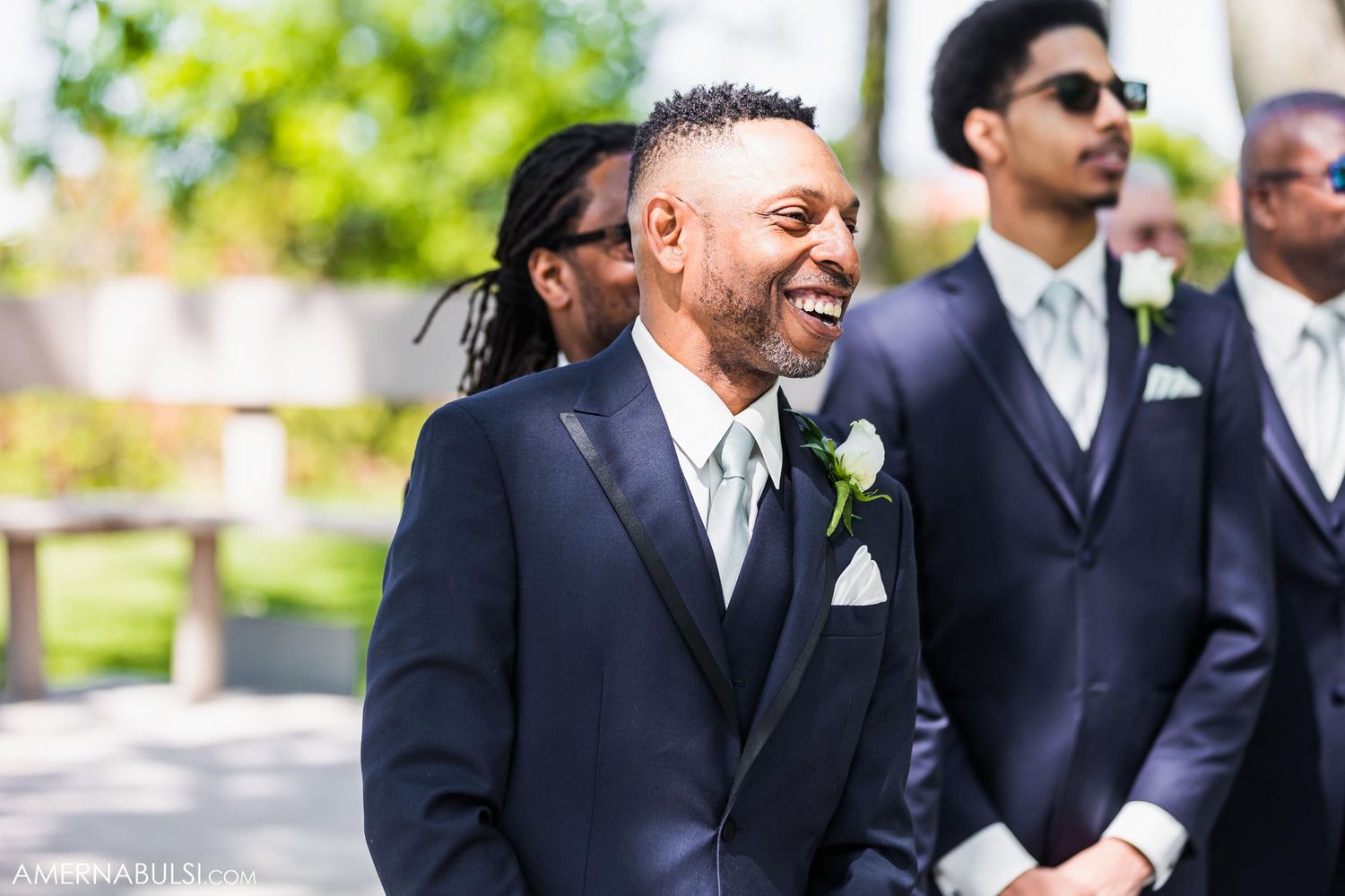 Groom in navy suit smiles with groomsmen lined up at outdoor ceremony.