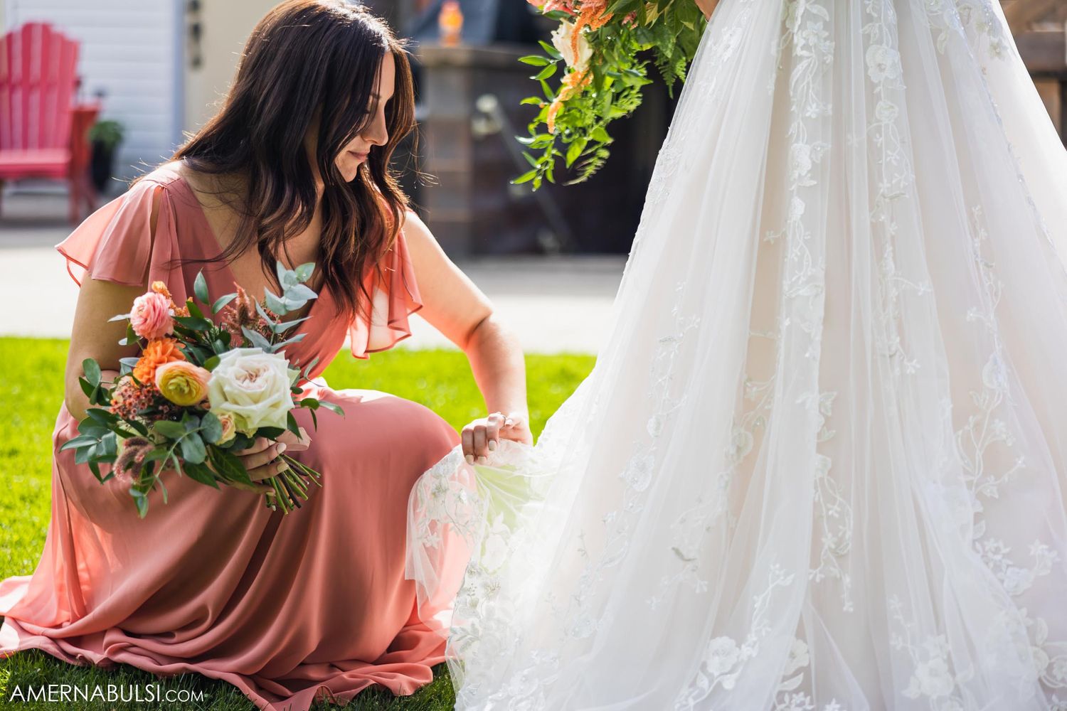 A bridesmaid in dusty rose dress arranges a wedding gown train on green grass.
