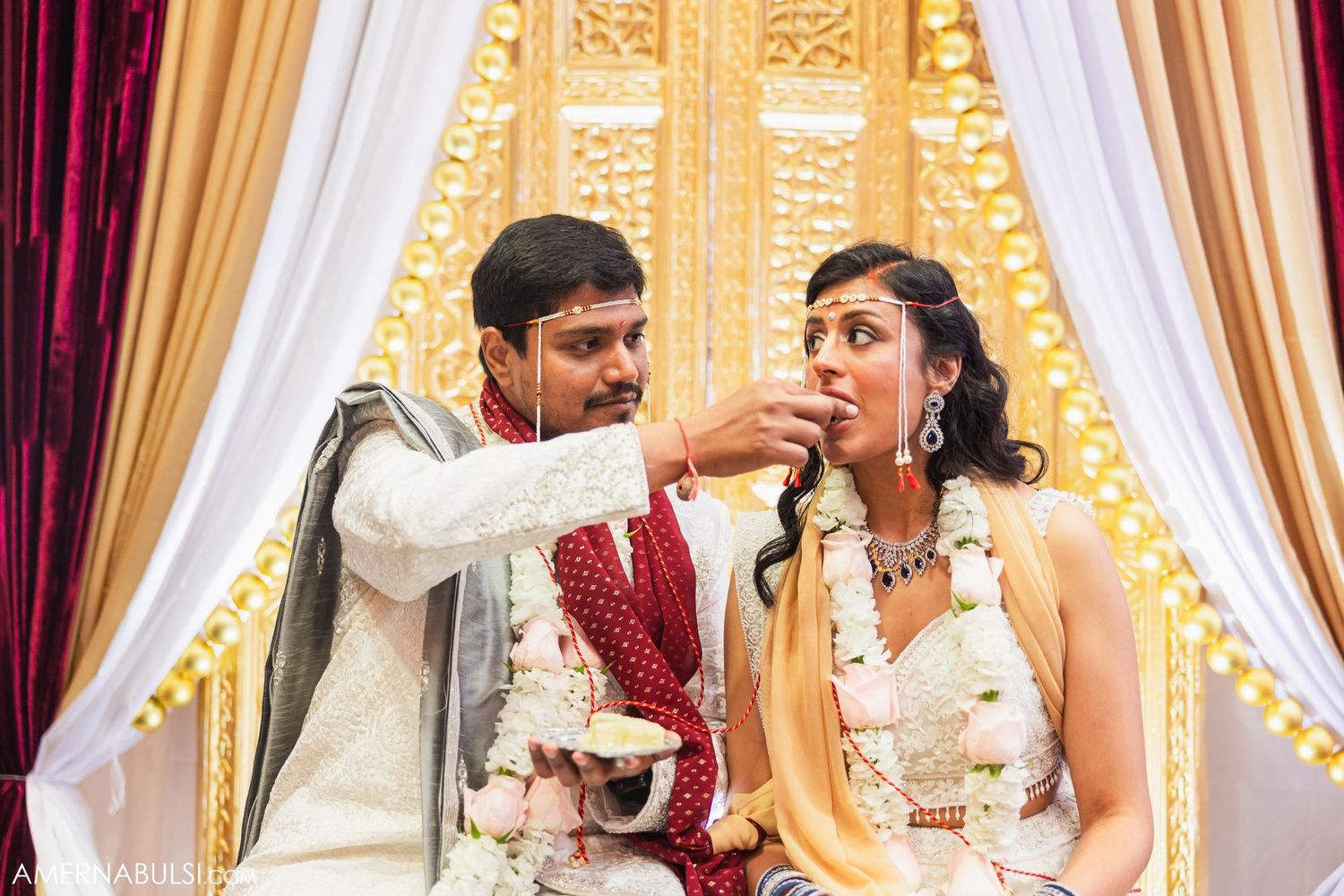 Couple participates in traditional Indian wedding feeding ritual during ceremony with decorative backdrop.