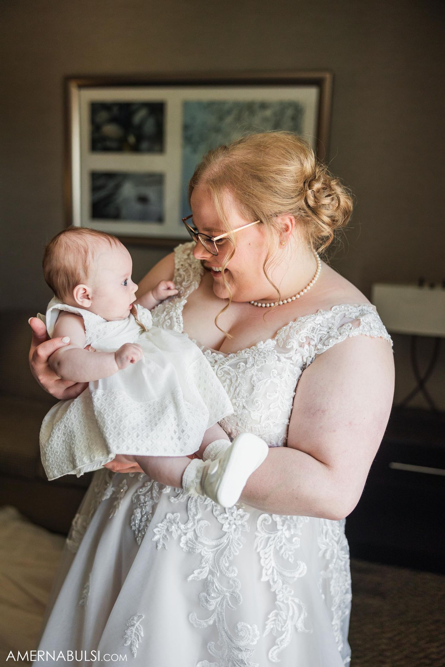Bride in white lace dress holding and smiling at baby in christening gown.