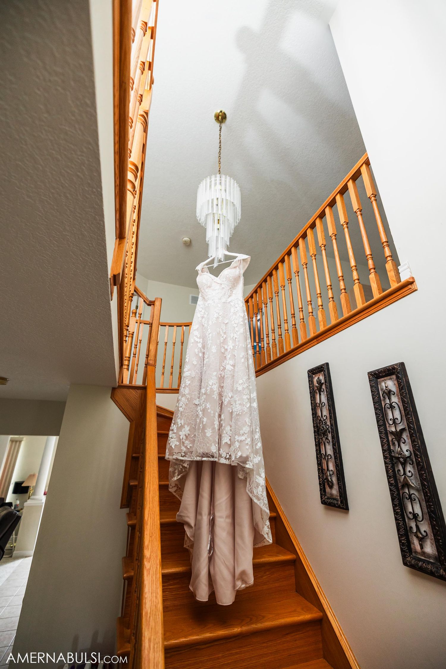 Elegant white wedding dress suspended from chandelier in staircase with wooden railing.