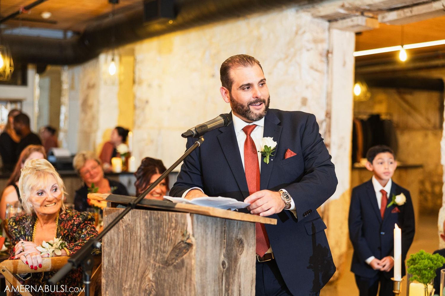 Speaker delivers wedding toast from wooden podium in industrial-style venue with exposed brick walls.