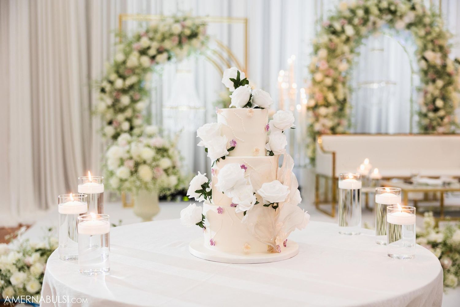 Elegant three-tiered white wedding cake decorated with flowers and surrounded by glowing candles on a round table.