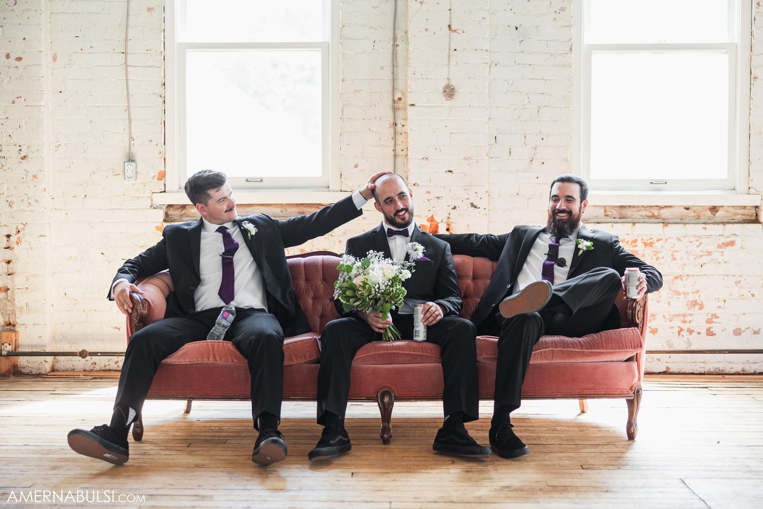 Wedding party relaxing and laughing on vintage red couch in industrial venue.