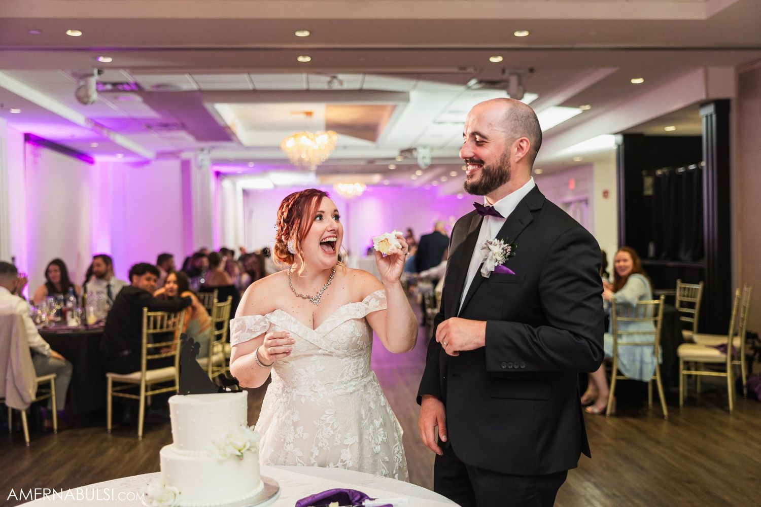 Bride and groom enjoying cake cutting moment at wedding reception with guests in background.