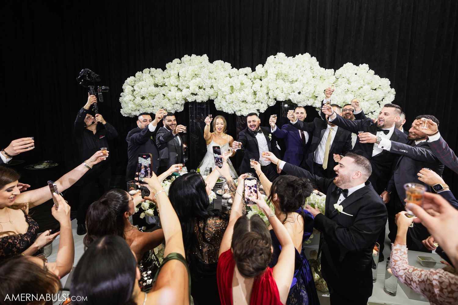 Wedding guests raise their phones to capture a celebration moment beneath elegant white floral arrangements.